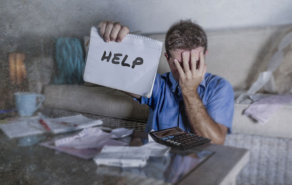 Man covers his face in shame while holding up a sign for help with his finances