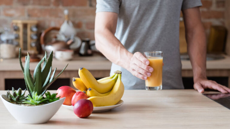 Through small changes, such as meal planning, we can save a lot of money. A man holds a glass of orange juice standing next to fresh fruit.